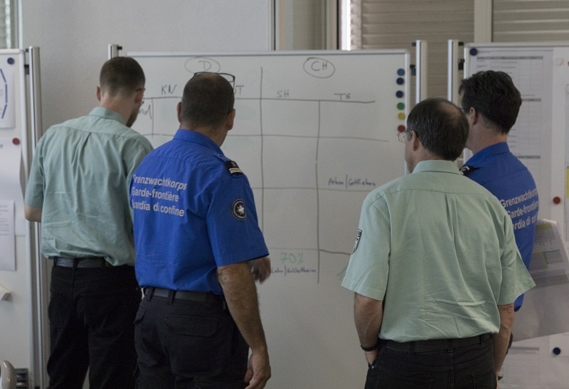 Zwei Einsatzleute des Grenzwachtkorps und zwei an der Übung teilnehmende Personen sehen sich Informationen auf einer Plakatwand an.