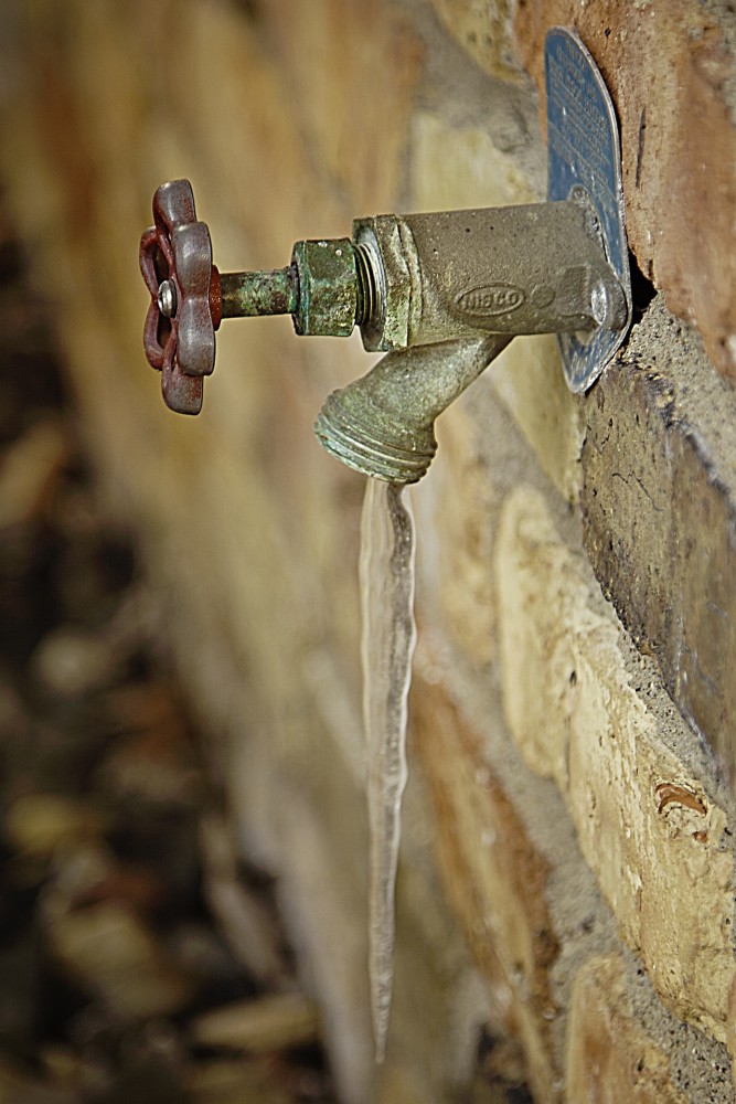 La photo montre un robinet fixé à un mur. L’eau a gelé pendant qu’elle s’écoulait du robinet, formant un glaçon. 